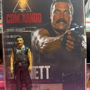 May include: A Commando action figure of the character Bennett in a plastic blister pack. The packaging features a photo of the character holding a gun, the text COMMANDO, BENNETT, and the quote I EAT GREEN BERETS FOR BREAKFAST. The figure wears a black leopard print vest.