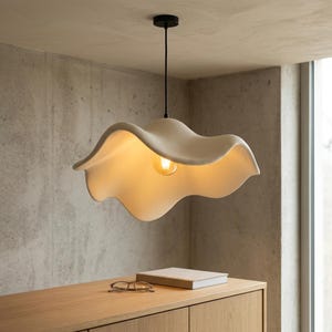 Może przedstawiać: Lampa wisząca z dużym, kremowobiałym kloszem o falistych, organicznych kształtach, zawieszona na cienkim czarnym przewodzie przy dopasowanej czarnej podsufitce. Wewnątrz klosza widoczna jest pojedyncza żarówka emitująca ciepłe światło.