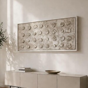 Pode incluir: Uma peça de arte de parede retangular com uma superfície bege texturizada e uma grade de elementos circulares em relevo, montada sobre um aparador longo de cor clara com quatro portas e pernas pretas finas em uma sala.