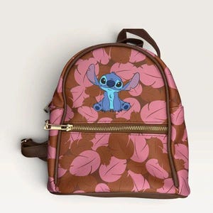 May include: A small backpack featuring a repeating pattern of pink and brown feathers. The center of the bag displays a blue Stitch character illustration. The backpack includes a front zippered pocket with gold-toned hardware and brown trim.
