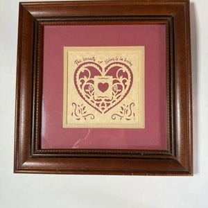 May include: A square dark brown wooden frame containing a maroon mat and a cream-colored paper cutout. The cutout features a heart-shaped filigree design with a central heart and the text, The beauty of living is in loving, printed above.