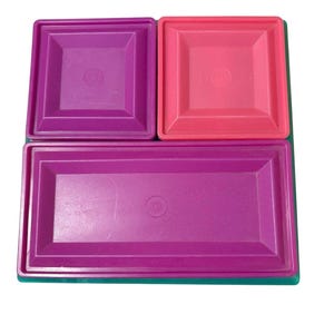 May include: A set of three plastic storage container lids in varying shapes and colours. There is one square purple lid, one square pink lid, and one large rectangular magenta lid. Each lid features a raised border and a central circular indentation.