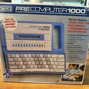 Może przedstawiać: Zabawka edukacyjna VTech PreComputer 1000 w oryginalnym niebiesko-białym pudełku. Opakowanie zawiera nazwę produktu, grafikę komputera z klawiaturą QWERTY i ekranem LCD oraz tekst opisujący funkcje takie jak nauka pisania i zadania matematyczne.