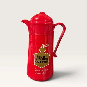 May include: A bright red thermal coffee carafe with a handle and a lid. The front features a gold-colored graphic of a coffee grinder with the text EIGHT O'CLOCK COFFEE and the words Quality Coffee Since 1869 printed in a script font below it.