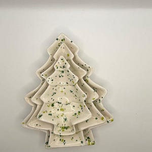 May include: A set of three nested ceramic dishes shaped like Christmas trees. Each dish is off-white with a speckled pattern of green and yellow dots. The dishes are stacked on top of each other, showing a consistent tree silhouette design.