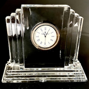 May include: A clear crystal desk clock with a silver-tone clock face and Roman numerals. The clock has a rectangular shape with Art Deco-inspired stepped sides. The clock face displays the "Waterford" brand.