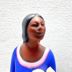 May include: A terracotta figurine of a person with dark skin and black hair, wearing a bright blue shirt with a pink collar. The figure holds a cluster of white calla lilies with yellow centers, rendered in a smooth, matte finish.