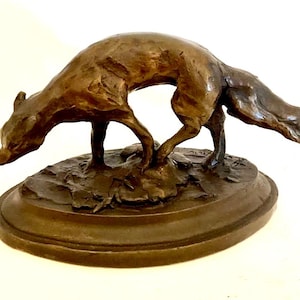 May include: Bronze fox sculpture in a hunting pose, with its head lowered and tail extended. The fox is mounted on an oval base with detailed foliage. The sculpture has a rich, dark brown patina, showcasing the intricate details of the animal.