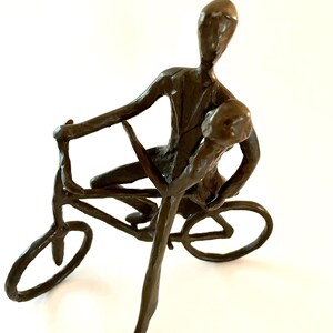 May include: Bronze sculpture depicting two figures on a bicycle. The figures are stylized, with elongated limbs and simplified features. The sculpture has a dark brown patina and is set against a white background.