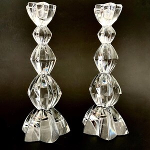 May include: Two clear crystal candle holders. Each holder has a faceted, geometric design with a flared base, a series of diamond-shaped segments, and a tulip-shaped top. The candle holders are displayed against a dark background.