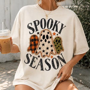 Puede incluir: Una camiseta de manga corta color hueso con el texto "SPOOKY SEASON" en letras negras desgastadas. El gráfico incluye tres ilustraciones de fantasmas: uno con un patrón de cuadros naranja y negro, uno con estrellas negras y uno con puntos verdes.