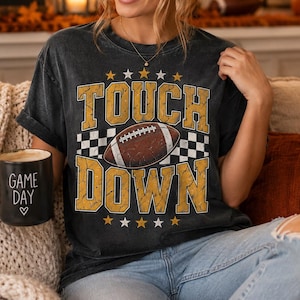 Puede incluir: Una camiseta de manga corta gris carbón con el texto TOUCH DOWN en letras amarillas desgastadas, un gráfico de balón de fútbol americano marrón y un patrón de cuadros blanco y negro. Incluye una taza negra con el texto GAME DAY y un corazón.