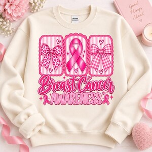 Op de afbeelding: Een crèmekleurige sweater met ronde hals en een grafische print met drie verticale panelen. De panelen tonen een roze strik met luipaardprint, een roze bewustzijnslint en een roze gingham strik. Daaronder staat de tekst: Breast Cancer AWARENESS.