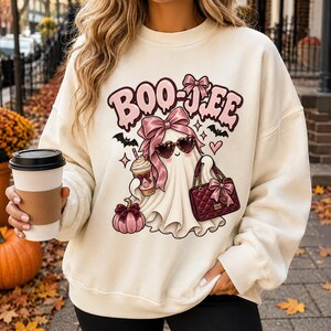 Puede incluir: Una sudadera de cuello redondo color crema con un gráfico de un fantasma que lleva gafas de sol en forma de corazón y un gran lazo rosa. El fantasma sostiene una taza de café y un bolso acolchado, con una calabaza y el texto BOO-JEE.