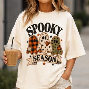 Puede incluir: Una camiseta de manga corta color blanco roto con un estampado gráfico de tres fantasmas y el texto negro SPOOKY SEASON. Los fantasmas tienen diferentes patrones, incluyendo cuadros naranjas y negros, blanco con estrellas negras y verde oliva con puntos negros.