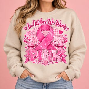 Op de afbeelding: Een beige sweater met ronde hals en een grafisch ontwerp met een grote roze bewustwordingslint, strikjes en hartjes. De tekst In October We Wear PINK staat in een vet, verweerd lettertype over een subtiel ruitpatroon op de voorzijde.