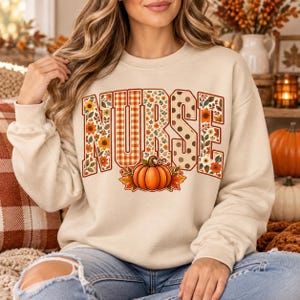 Puede incluir: Una sudadera beige de cuello redondo con la palabra NURSE en letras grandes. Las letras están rellenas con patrones de girasoles naranjas, hojas de otoño, cuadros vichy naranjas y lunares marrones, con una calabaza naranja en el centro.