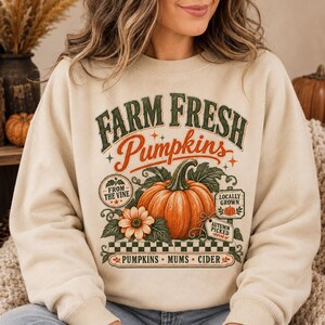 Puede incluir: Una sudadera beige de cuello redondo con un estampado gráfico que incluye el texto "FARM FRESH Pumpkins". El diseño muestra una calabaza naranja grande, una flor y pequeños letreros que dicen "FROM THE VINE", "LOCALLY GROWN" y "AUTUMN PICKED".