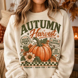 Puede incluir: Una sudadera color crema con un estampado gráfico que incluye el texto AUTUMN Harvest. El diseño muestra una calabaza naranja grande, un girasol, hojas y letreros que dicen PATCH GROWN, OPEN WEEKENDS, COZY SEASON, además de PUMPKINS BAKED PIES HOT CIDER.