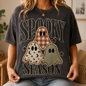 Puede incluir: Una camiseta gris carbón de manga corta con el texto SPOOKY SEASON en una fuente negra desgastada. El diseño incluye tres ilustraciones de fantasmas con bordes cosidos: uno de cuadros marrones, uno crema con estrellas y lunas negras, y uno de cuadros verdes.