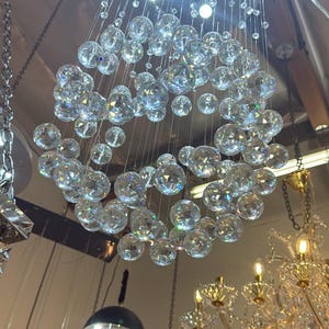 May include: A circular chandelier featuring numerous clear, faceted crystal spheres suspended at varying lengths from a round ceiling mount. The light reflects through the glass orbs, creating a bright, multi-layered display of illumination.