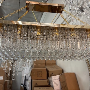 May include: A rectangular chandelier featuring a polished gold-tone metal frame and multiple tiers of clear, faceted crystal prisms and hanging crystal beads. The fixture is suspended by gold-tone chains attached to a matching rectangular canopy.