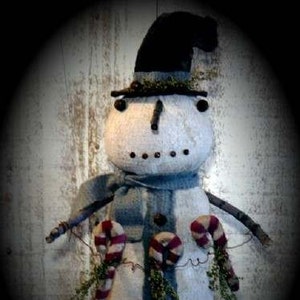 Pode incluir: Um boneco de neve primitivo feito à mão com um chapéu preto, um cachecol listrado azul e branco e bengalas de doces. O boneco de neve é feito de tecido branco e tem um visual rústico e vintage.