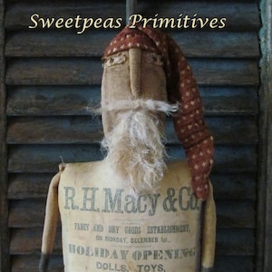 May include: A handmade rag doll dressed as Santa Claus. The doll is made from burlap fabric and has a white beard. The doll is wearing a red and white stocking cap and is holding a red bauble. The doll is standing in front of a wooden background. The doll is advertising a holiday opening for R.H. Macy & Co. on December 1st. The ad includes the text "Holiday Opening", "Dolls, Toys, Fancy Goods, Christmas Gifts", and "Doll Life."