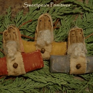 May include: Three handmade primitive Santa Claus ornaments. Each ornament features a tan head with a white beard, a bell, and a colorful base in red, yellow, and blue. The ornaments are displayed on a bed of green foliage, with the text "Sweetpea's Primitives" in the top left corner.