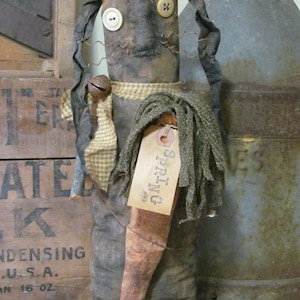 以下が含まれることがあります： 茶色の布製の体と黒い布製の髪、大きな白いボタンの目をした手作りのかやの人形。人形は茶色のニンジンと「春」と書かれたタグを持っています。