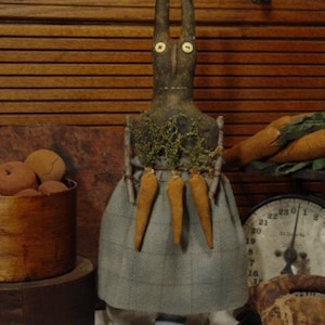 May include: A handmade primitive style bunny doll with long ears, wearing a blue and white plaid skirt and holding a bunch of carrots. The doll is standing on a glass jar with a blue base.