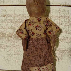 Könnte beinhalten: Handgefertigte Puppe mit braunem Kopf und Körper, trägt ein gemustertes Kleid. Das Kleid hat ein Blumenmuster mit roten Beeren und das Jahr "1873" auf der Vorderseite aufgenäht. Die Puppe hat eine rustikale, Vintage-Ästhetik.