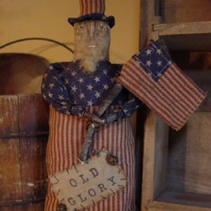 May include: A patriotic doll figure with a top hat and an American flag. The doll is dressed in red and white striped fabric with a blue star-patterned shirt. A sign reading "OLD GLORY" hangs from its twig arms.