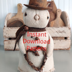 Peut inclure: Un bonhomme de neige en tissu blanc portant un chapeau en feutre marron et une écharpe marron avec des clochettes. Le bonhomme de neige tient une couronne de brindilles en forme de cœur. Le texte "Instant Download Pattern" est imprimé sur la poitrine du bonhomme de neige.