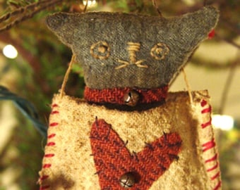 Op de afbeelding: Handgemaakte kerstversiering in de vorm van een kat. De grijze kattenkop heeft geborduurde ogen en snorharen. Het beige lichaam heeft een rood hart en is afgezet met rode stiksels. Een klein belletje hangt aan de nek.