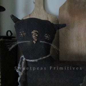 May include: Black fabric cat with a stitched face and whiskers. The cat is wearing a brown fabric tie and the text "Sweetpeas Primitives" is visible on the bottom of the image.