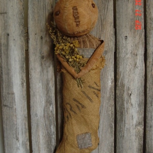 May include: A burlap stocking with a stitched face and a twig nose. The stocking is holding a bouquet of dried flowers. The stocking is hanging on a wooden fence.