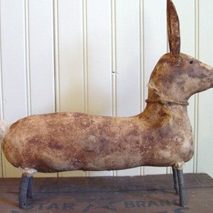 Puede incluir: Un conejo de estilo primitivo, hecho a mano, de color marrón con patas de alambre. El conejo tiene un cuerpo largo y delgado y una cola larga y delgada. El conejo está hecho de tela y tiene un aspecto rústico y desgastado.
