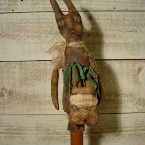 May include: A rustic, brown, handmade bunny doll with long ears and a burlap sack that says "Carrots" hanging from its side. The bunny is holding a bunch of green fabric carrots in its hands. The bunny is standing on a wooden base.