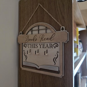 May include: A light-coloured wooden wall hanging sign with a hanging chain. The sign features an arched top, the text "Books Read THIS YEAR" in black, three small hooks for display, and a graphic of an open book with decorative swirls.