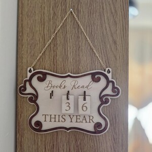 May include: A light-coloured wooden wall plaque with a dark brown decorative border and a gold chain for hanging. The text reads Books Read THIS YEAR, featuring two small wooden tiles displaying the numbers 3 and 6 hanging from metal hooks.