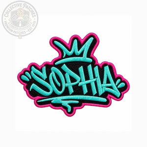 May include: An embroidered patch featuring the name SOPHIA in bright turquoise graffiti-style lettering with a black background and a vibrant hot pink border. Above the name is a stylized turquoise crown, all outlined in hot pink thread.