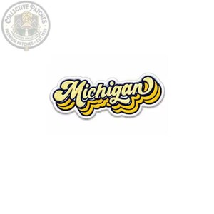 May include: An embroidered patch featuring the word Michigan in a stylized, cursive font. The lettering is yellow with a dark blue outline and a white border, creating a layered, wavy effect. The design is isolated against a plain white background.