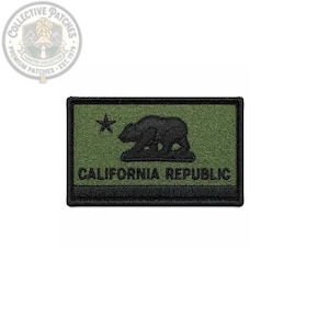 Puede incluir: Un parche rectangular bordado con fondo verde oliva y borde negro. El diseño incluye una estrella negra, la silueta de un oso negro y el texto CALIFORNIA REPUBLIC en letras mayúsculas negras centrado debajo del oso.
