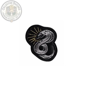 May include: An embroidered patch featuring a silver coiled snake on a black background. Behind the snake is a gold sunburst design with radiating lines. The patch has a black border and an irregular, rounded shape with detailed stitching.