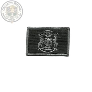 Könnte beinhalten: Ein rechteckiger, schwarz bestickter Aufnäher mit einem einfarbigen grauen Design des Wappens des Bundesstaates Michigan. Das Emblem zeigt zwei Hirsche, die auf beiden Seiten eines Schildes stehen, mit dem lateinischen Wort TUEBOR in der Mitte.