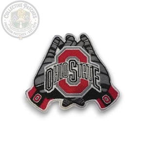 Puede incluir: Un parche bordado que muestra dos guantes de fútbol americano cruzados en gris y negro con detalles rojos en las muñecas. Centrado sobre los guantes hay un logotipo rojo y blanco de Ohio State con el texto OHIO STATE en letras negras.