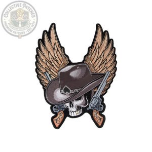 May include: An embroidered patch featuring a skull wearing a brown cowboy hat with a black spade emblem. Two crossed revolvers with ornate grips are positioned behind the skull, and large, detailed tan wings extend upward from behind the hat.