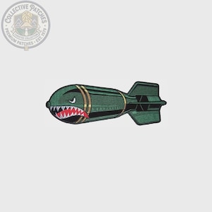 May include: An embroidered patch shaped like a green bomb with a shark mouth graphic featuring white teeth and a red tongue. The design includes two gold bands around the body and a finned tail section, rendered in dark green thread with black outlines.
