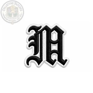 May include: An embroidered patch featuring the letter M in a black Old English font with a thick white border. The design is centered on a plain background, showcasing detailed stitching and a high-contrast color scheme for clear visibility.
